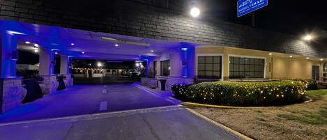 Entrada de um hotel com área de estacionamento coberta, uma placa indicando 'Hotel Away from Home' e um caminho bem iluminado que leva à entrada.