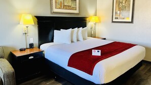 Una cama bien hecha con sábanas blancas y una manta roja, flanqueada por dos mesitas de noche con lámparas, en una habitación con cuadros en las paredes.