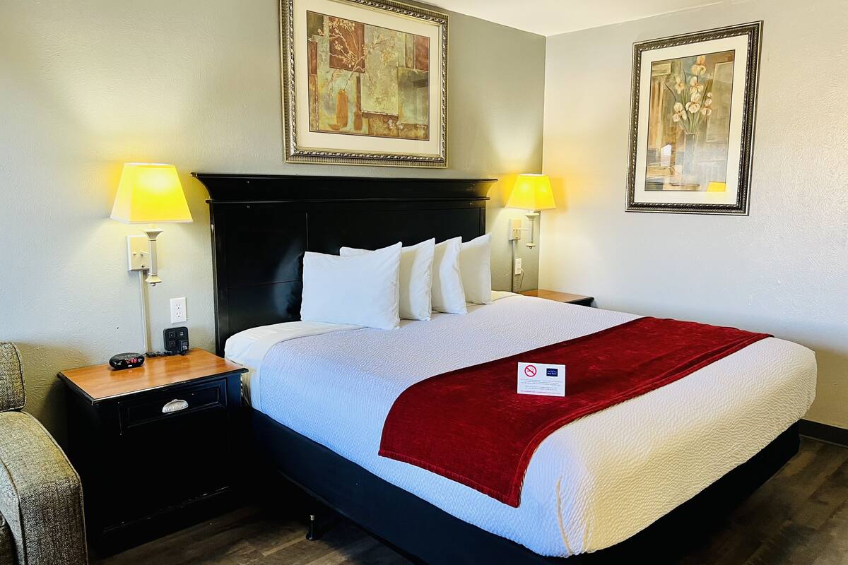 A neatly made bed with white linens and a red throw, flanked by two bedside tables with lamps, in a room with framed artwork on the walls.