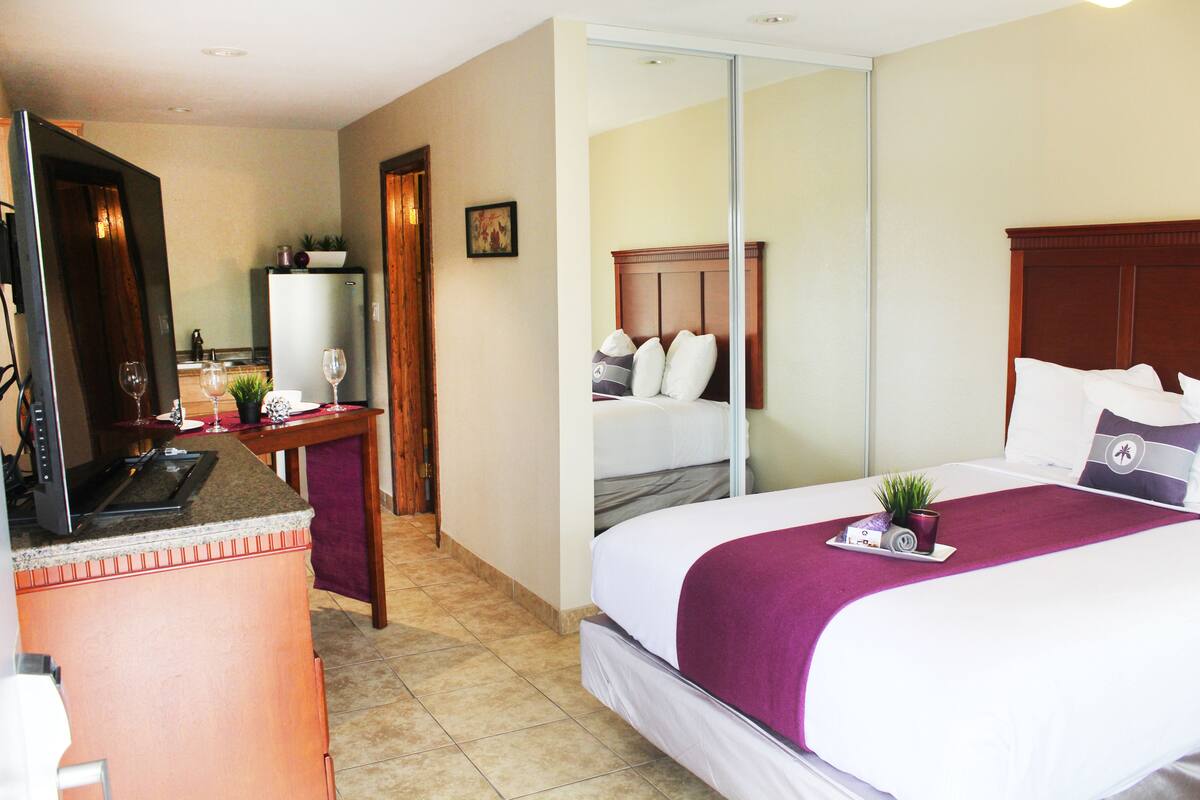 Una habitación de hotel con cama, escritorio con computadora, refrigerador y espejo.