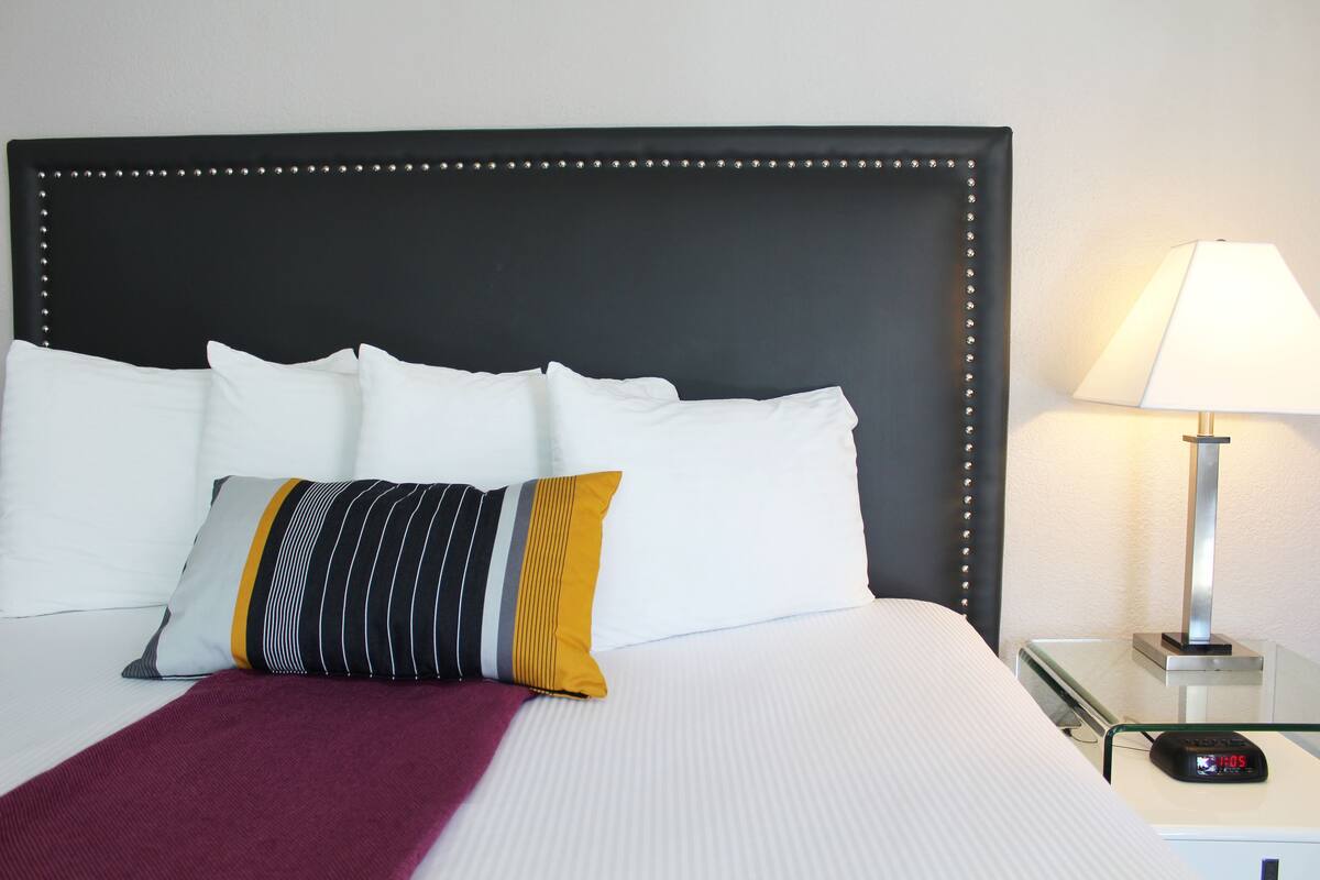 Una cama bien hecha con sábanas blancas, un cabecero negro adornado con tachuelas y una mesita de noche con una lámpara y un reloj digital.