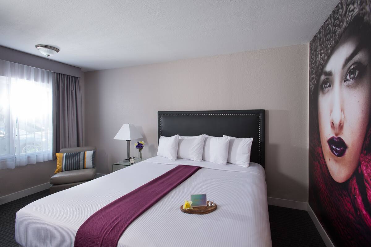 Habitación de hotel con una cama grande, cabecera, mesita de noche, lámpara, silla, ventana con cortinas y una obra de arte colgada en la pared.