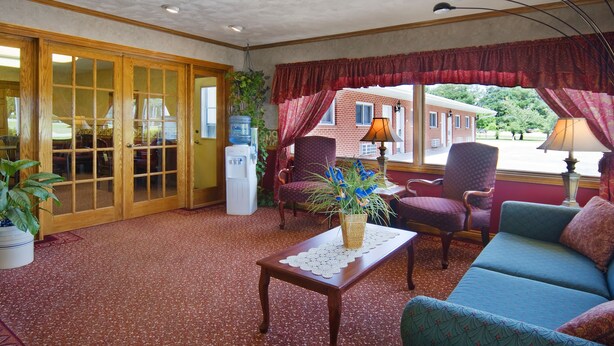 A room with a red carpet, wooden-framed sliding doors, a blue sofa, a patterned armchair, a small table with a doily, and a window with red curtains. - Americas Best Value Inn York (York)