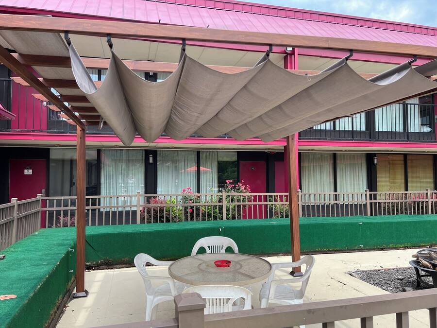 A covered outdoor seating area with a table and chairs, a green lawn, and a building with pink and black exterior walls.