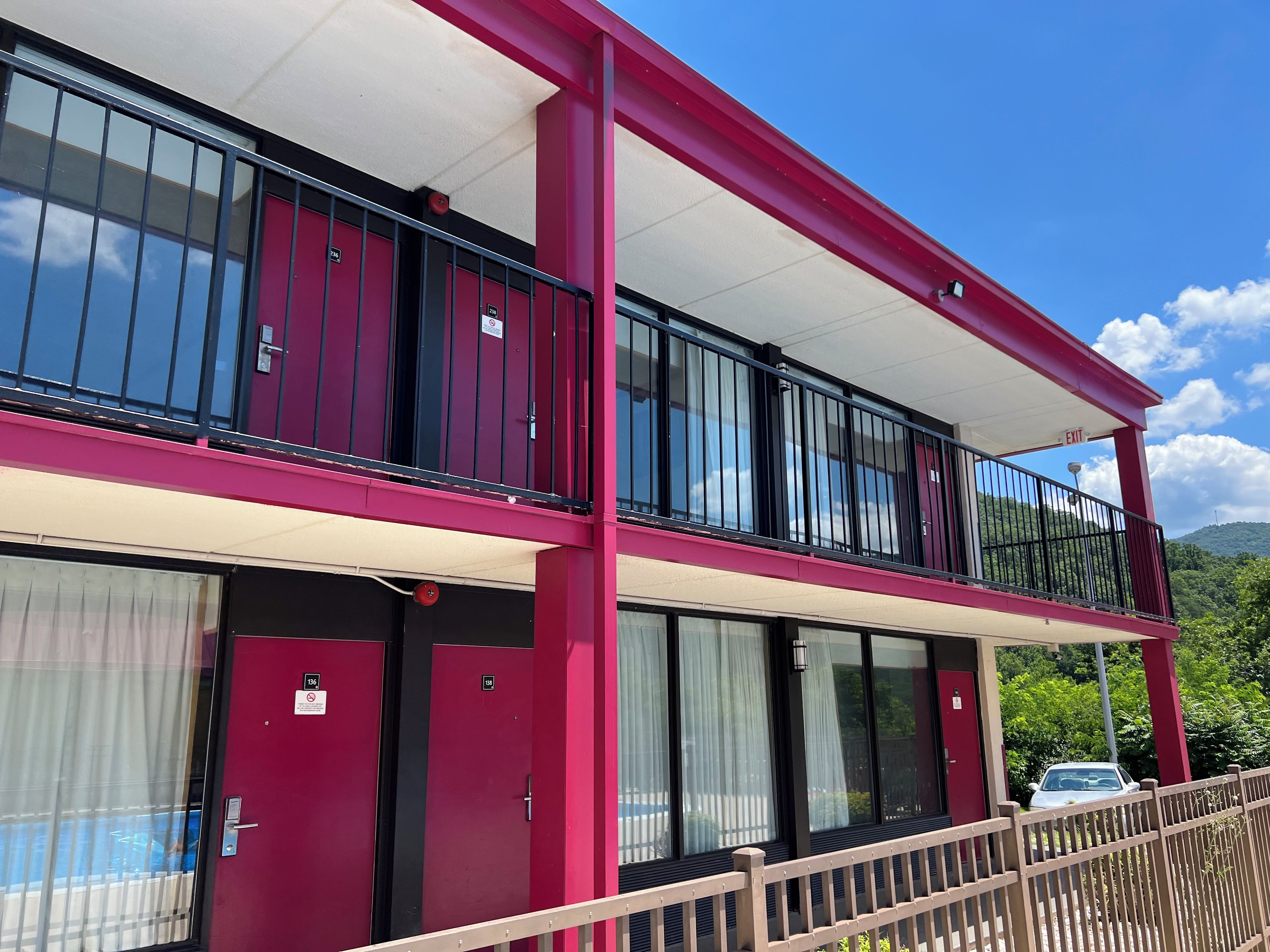 A two-story building with a red exterior, balconies with black railings, and a pool area visible through a glass door.