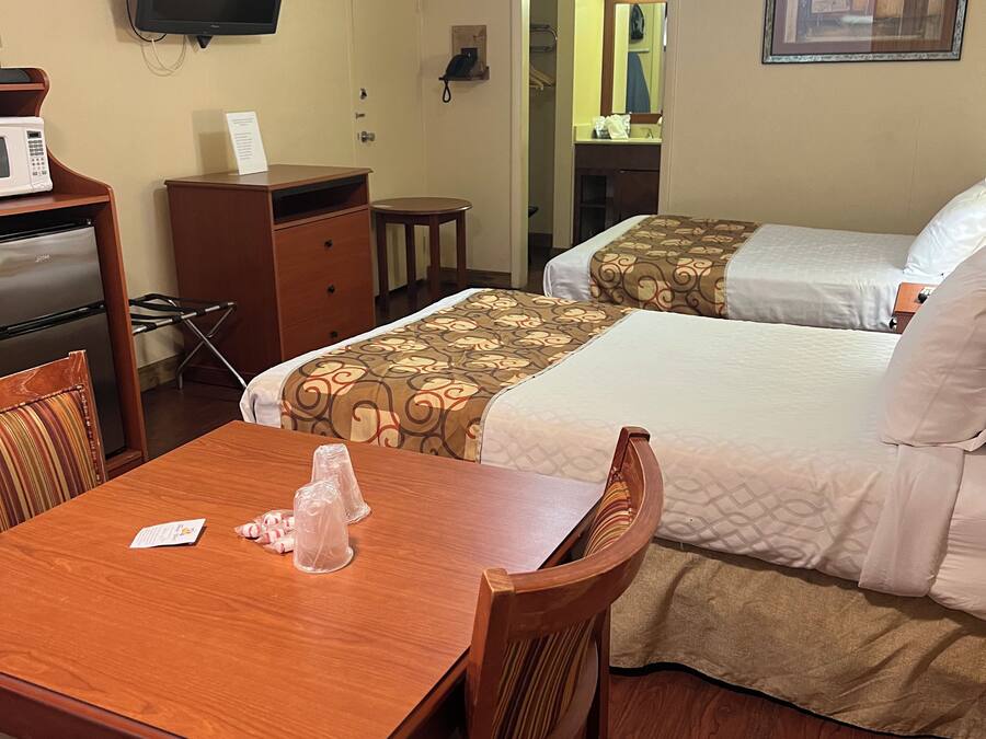 A hotel room with a wooden table, two beds, a ceiling fan, a television, and a microwave.