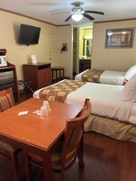 Habitación de hotel con una mesa de madera, dos camas, un ventilador de techo, un televisor y un microondas.