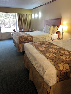 A hotel room with two beds, a nightstand, a lamp, and a window with curtains. - Red Rose Hotel (Covington)