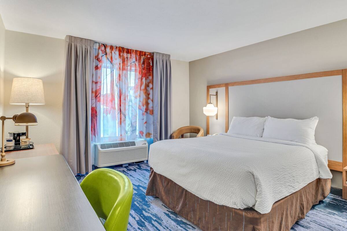 A hotel room with a large bed, a desk with a lamp, a chair, and a window with curtains.