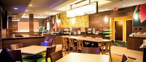 A hotel restaurant with multiple dining tables and chairs, a buffet area, and a kitchen in the background.
