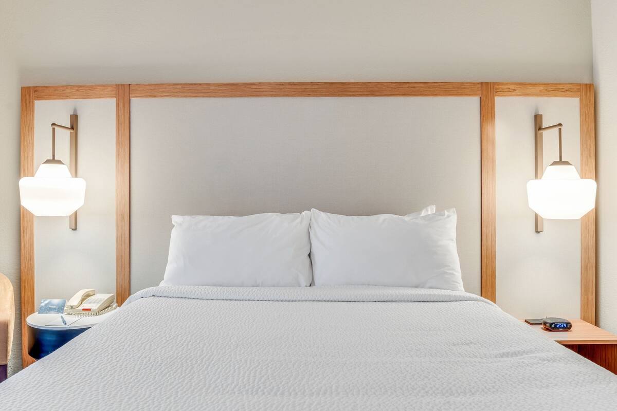 A neatly made bed with white bedding, a wooden headboard, bedside lamps, a bedside phone, and a digital clock.