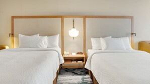 Una camera d'albergo con due letti, una testiera in legno, un comodino con una lampada e un tavolino con una sveglia.