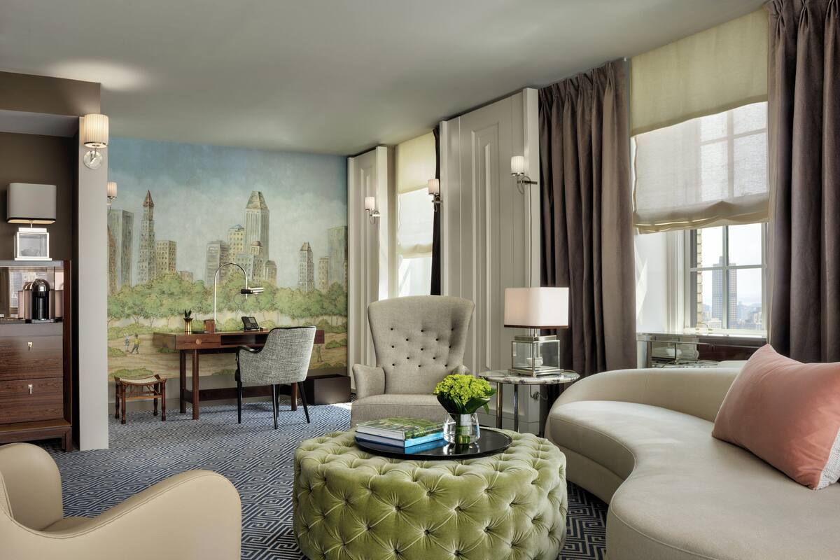 A hotel room with a cityscape mural, a green ottoman, a sofa, a desk with a chair, and a window with curtains.