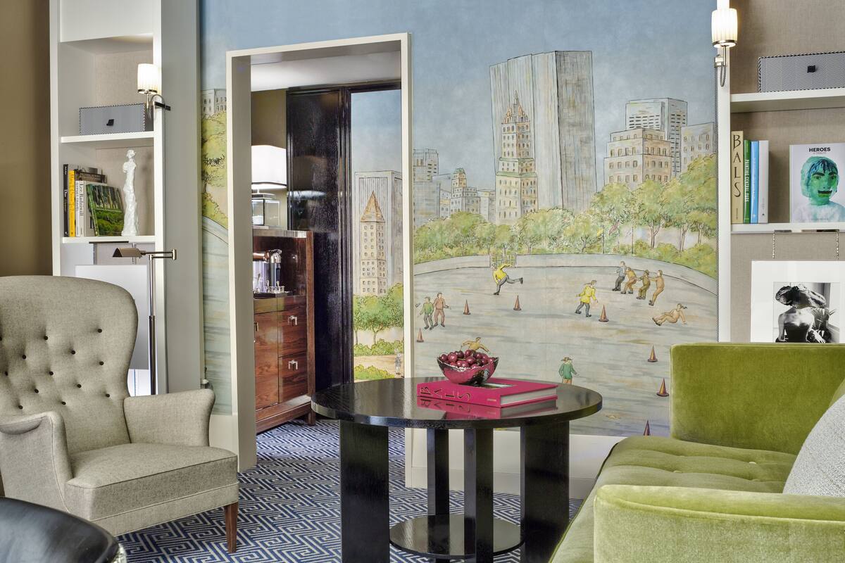 A living room with a cityscape mural, a round black table, a green armchair, and a beige armchair.