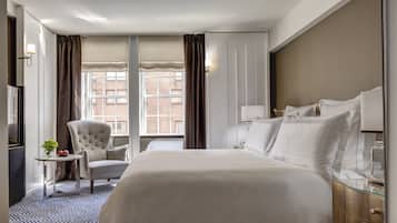 A hotel room with a large bed, a chair, a small table with fruit, and a window with curtains.