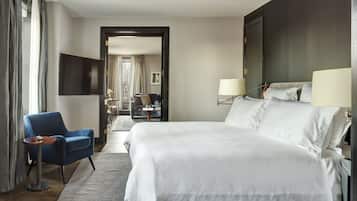 A hotel room with a large bed, a blue armchair, a small table, a TV, and a view of the city through the window.