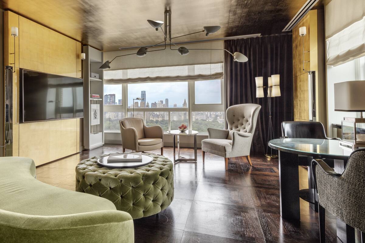 A modern hotel room with a large window, a flat-screen TV, a round glass table, and a tufted ottoman.