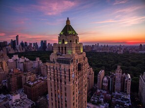 A high-rise building with a distinctive green-lit spire, surrounded by dense urban skyscrapers and a city skyline at sunset.