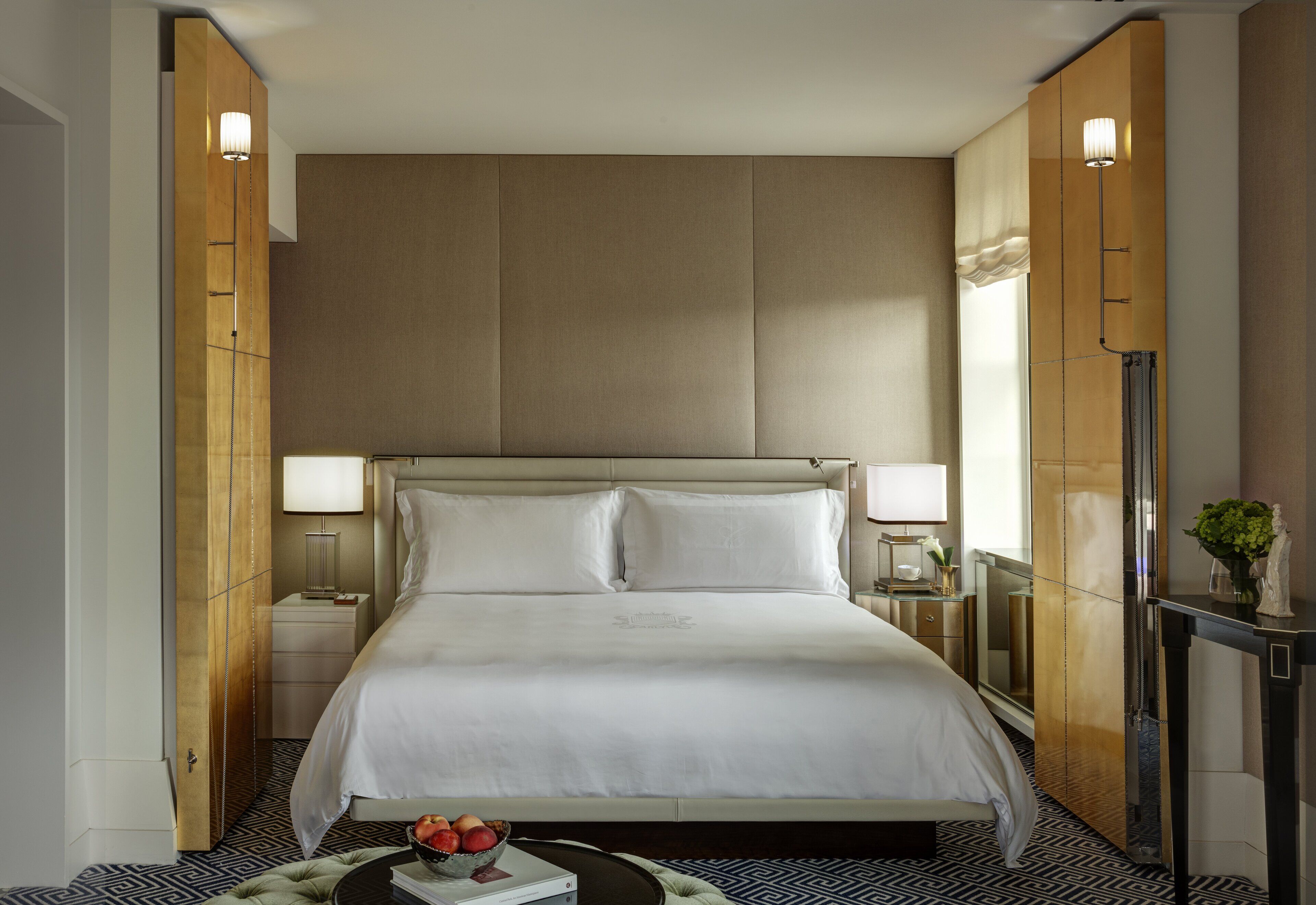 Een hotelkamer met een groot bed, twee bedlampen, een nachtkastje, een klein tafeltje met een vaas en een kamer met een patroon op de vloer.