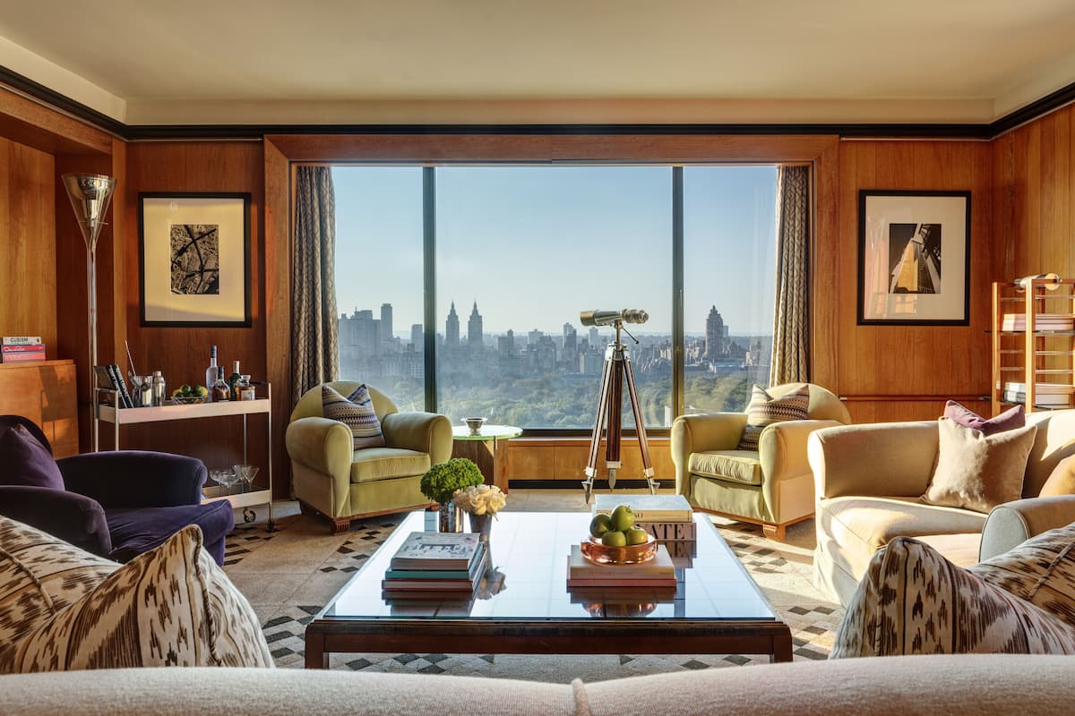 A spacious living room with a large window offering a city view, two sofas, a coffee table with books and decorative items, and a telescope on the windowsill.