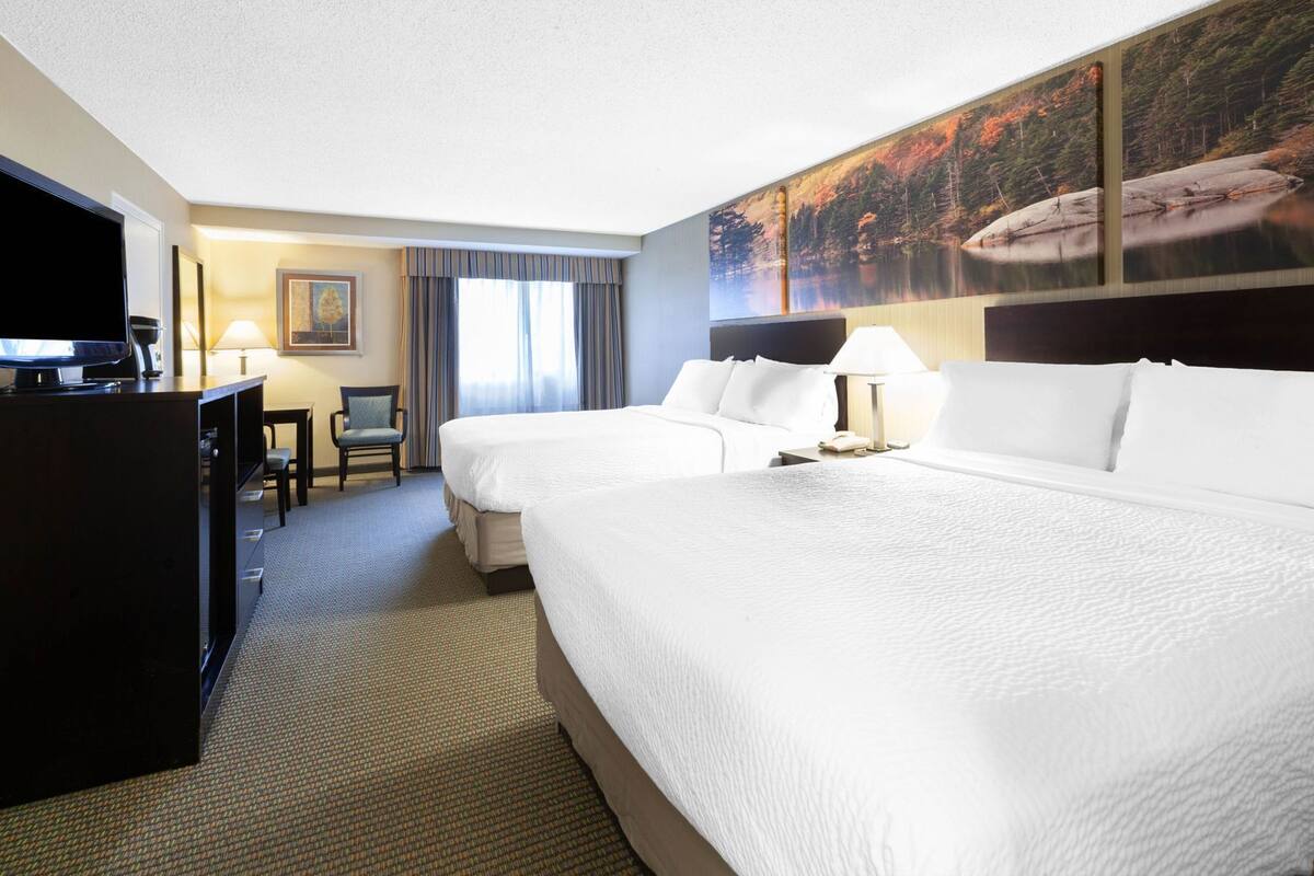 A hotel room with two beds, a desk, a TV, and a painting of a forest scene.