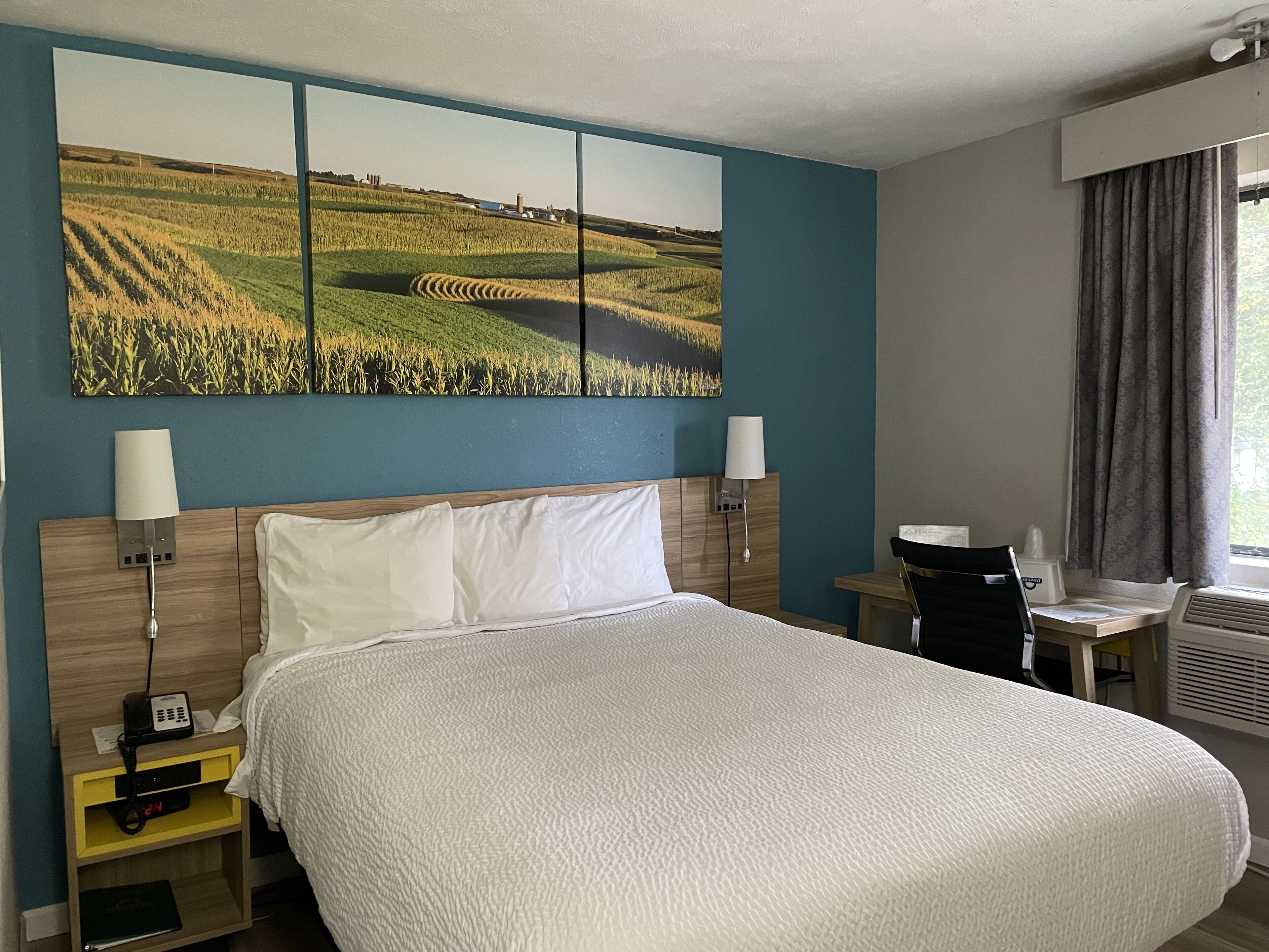 A hotel room with a large bed, two bedside lamps, a desk with a chair, and a large landscape painting on the wall.