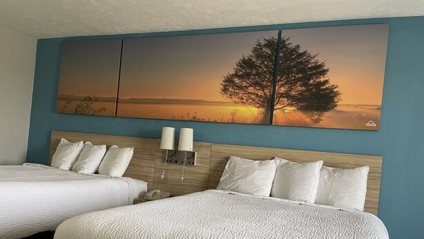 Habitación de hotel con dos camas, un cuadro grande en la pared con un árbol al atardecer y dos lámparas de pared.