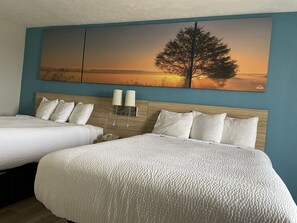 A hotel room with two beds, a large wall painting of a tree at sunset, and two wall-mounted lamps.
