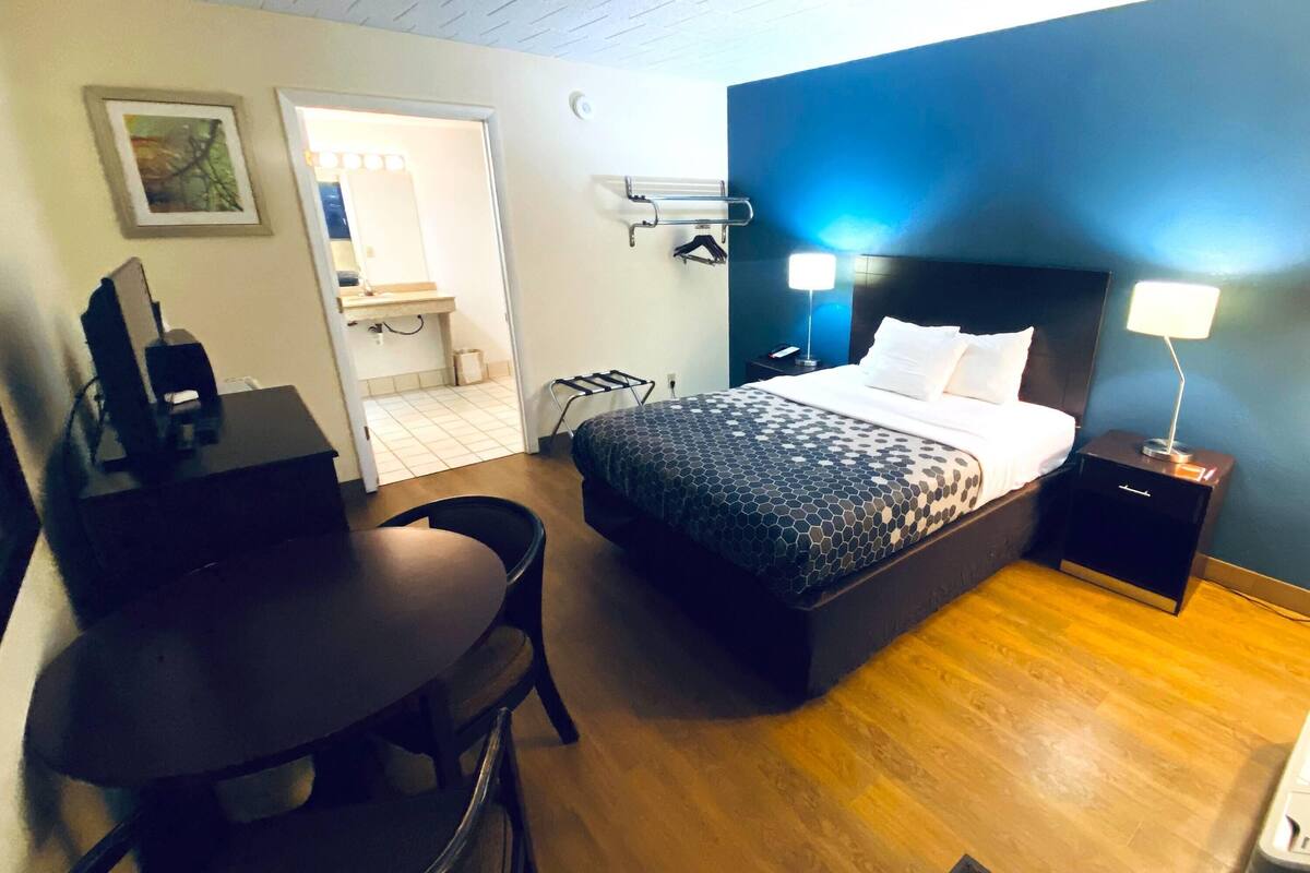 Une chambre d’hôtel avec un lit, une table de chevet, un bureau, une chaise et une salle de bain visible à travers une porte ouverte.