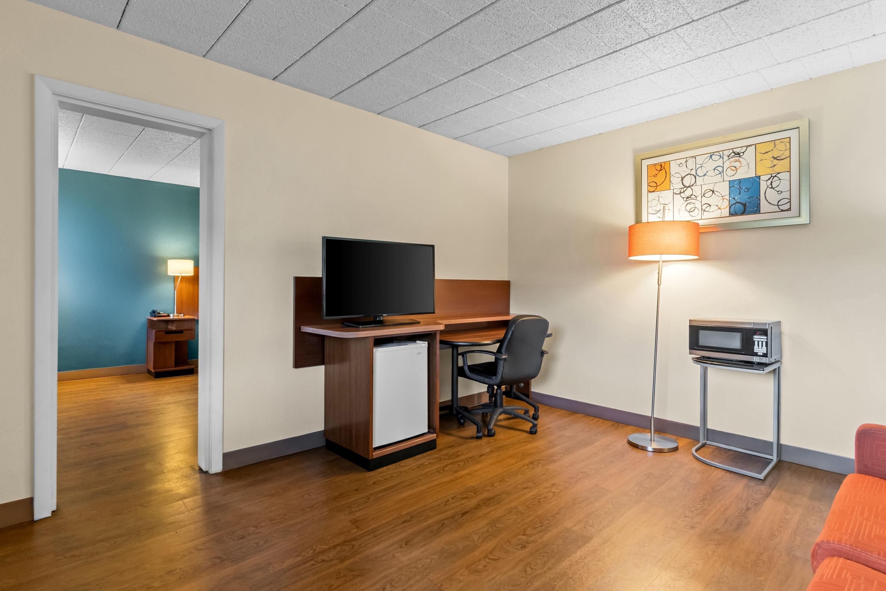 A hotel room with a wooden desk, a television, a small refrigerator, a desk lamp, a microwave, and a framed abstract art piece on the wall.