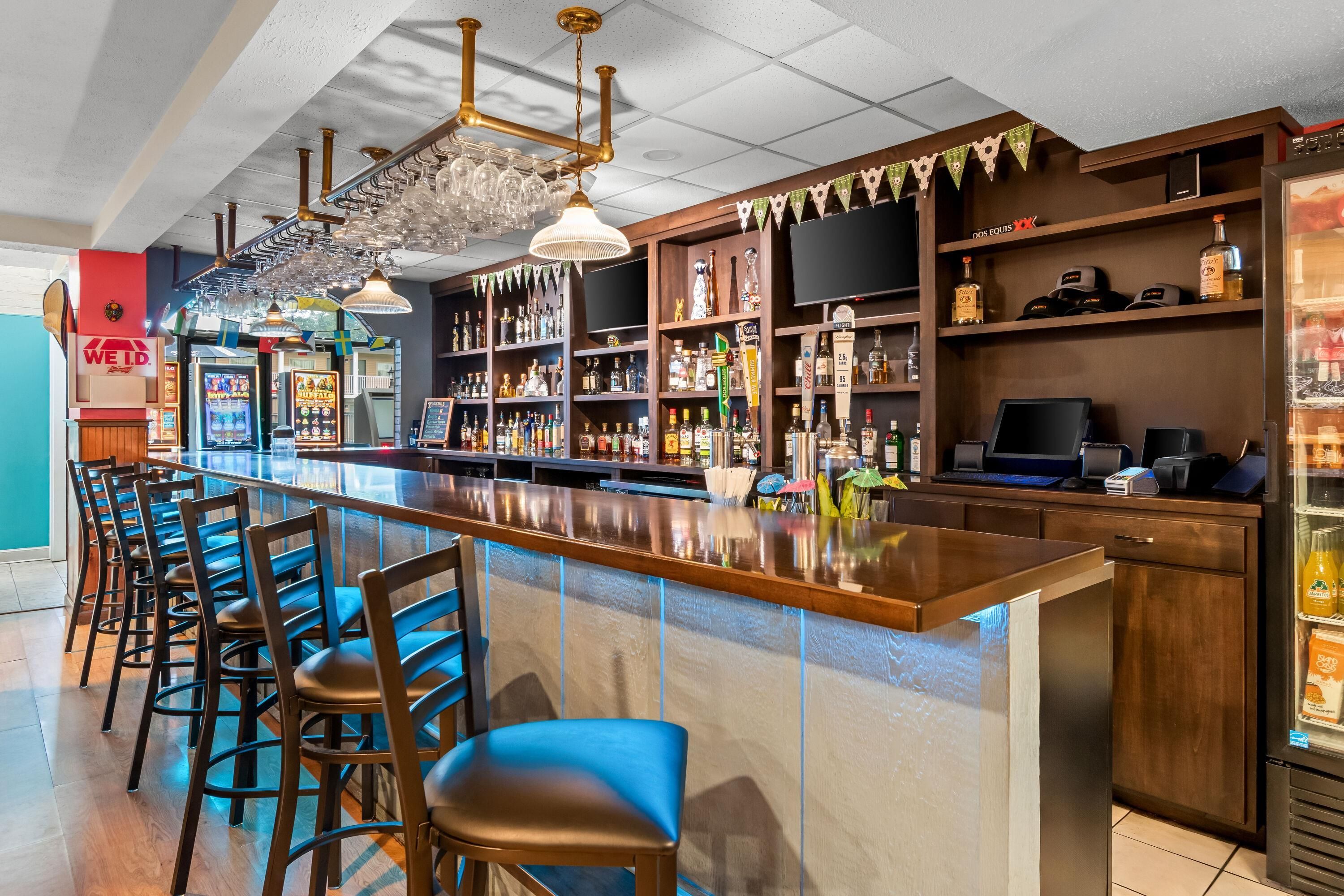 A bar area with a long counter, multiple stools, and a variety of bottles and glasses on shelves.