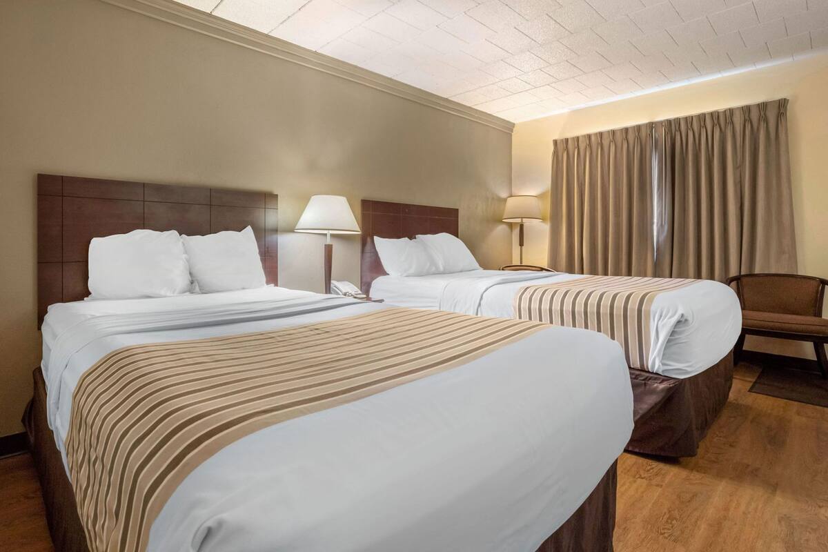 Une chambre d’hôtel avec deux lits, une tête de lit en bois, une table de chevet avec une lampe, une chaise et une fenêtre avec des rideaux.
