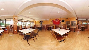 Een ruime eetruimte met meerdere tafels en stoelen, een grote klok aan de muur en uitzicht op de omgeving door grote ramen.
