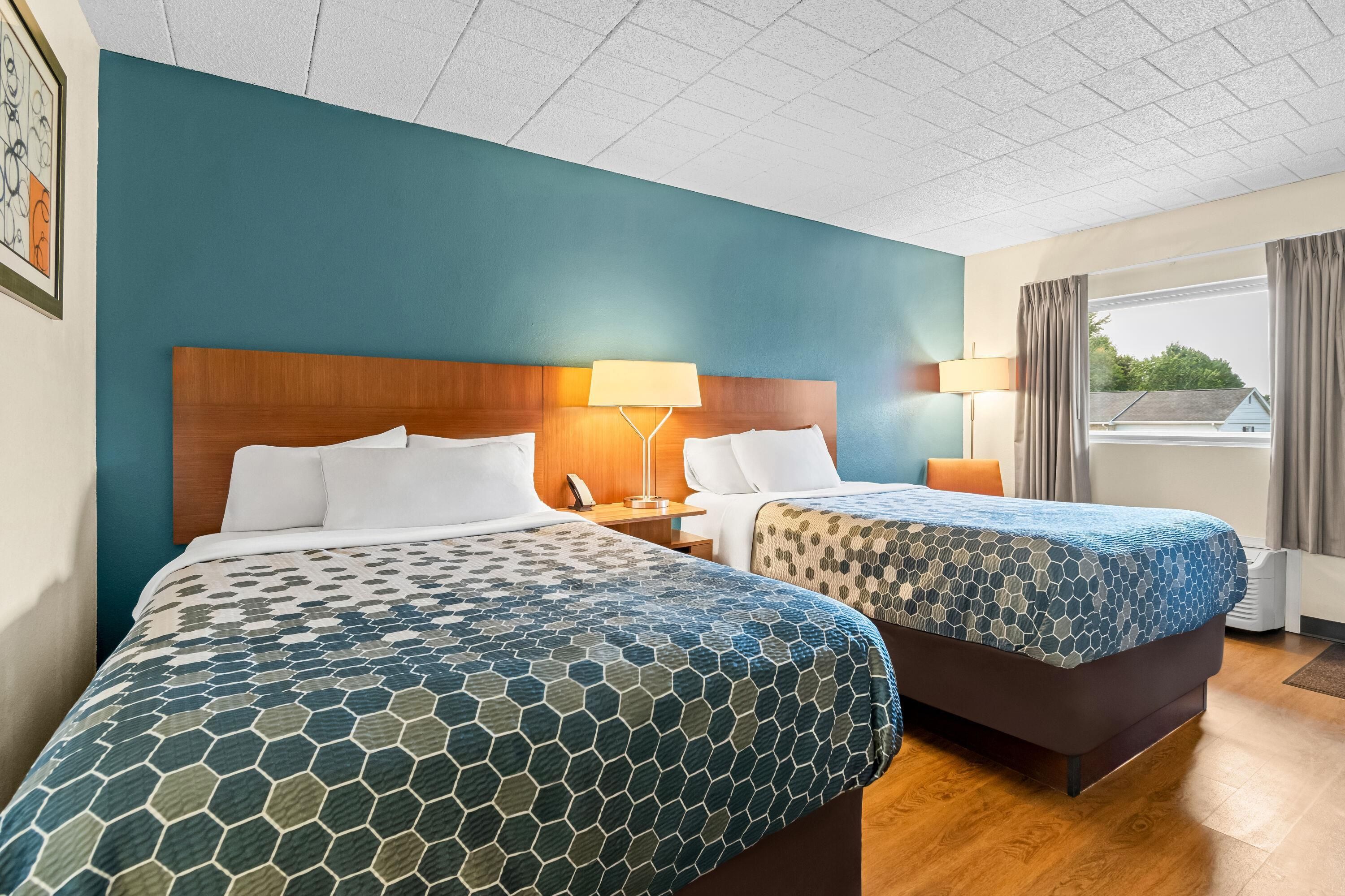 Habitación de hotel con dos camas, cabecera de madera, mesita de noche, lámpara y vistas al exterior.