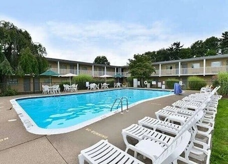 A swimming pool with lounge chairs and umbrellas, surrounded by a building with a flat roof and a fence.. Travelodge by Wyndham Shamokin Dam/Selinsgrove