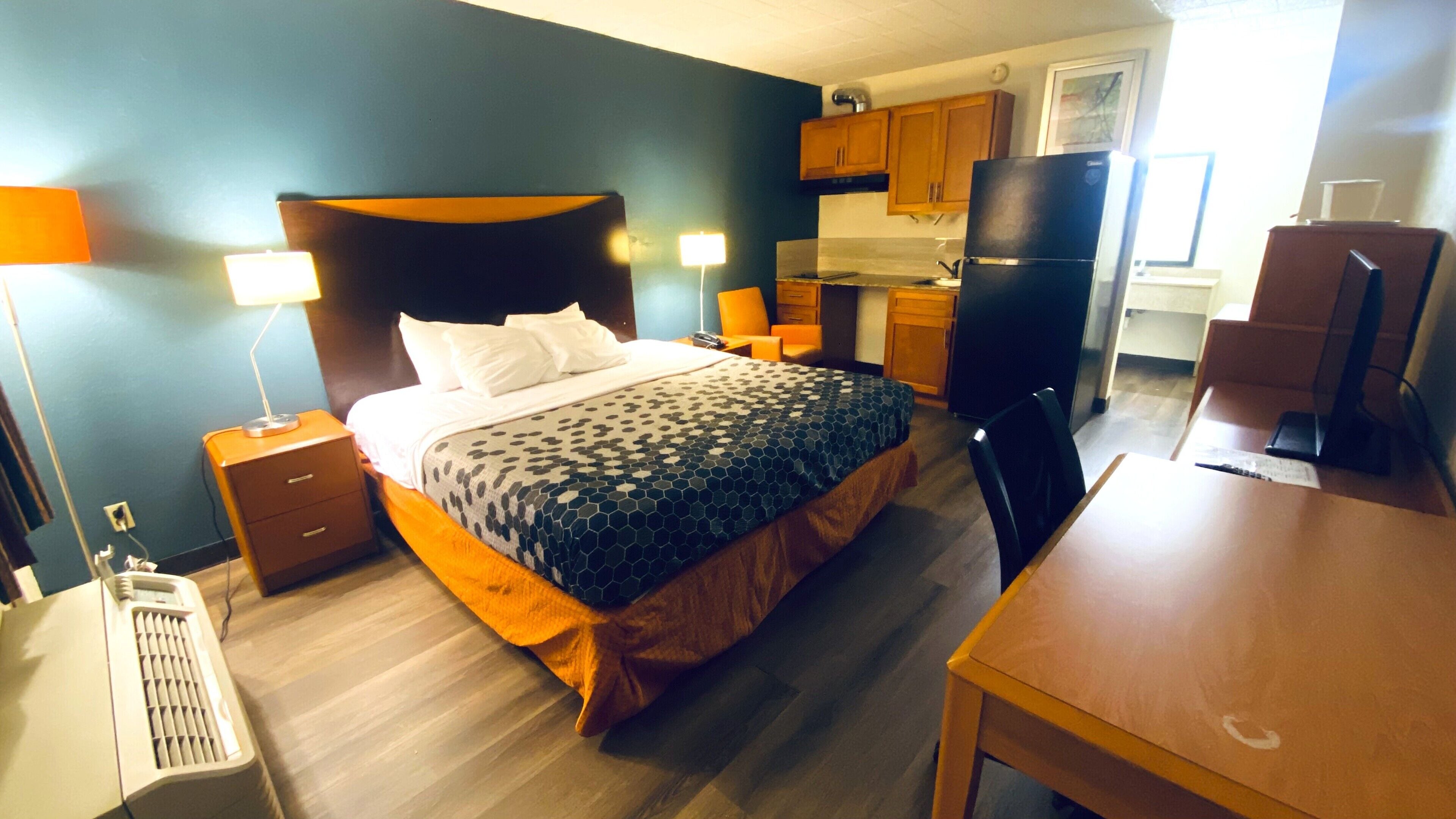 Quarto de hotel com cama, mesa de cabeceira, luminária, ar condicionado e uma área de cozinha compacta com geladeira e fogão.