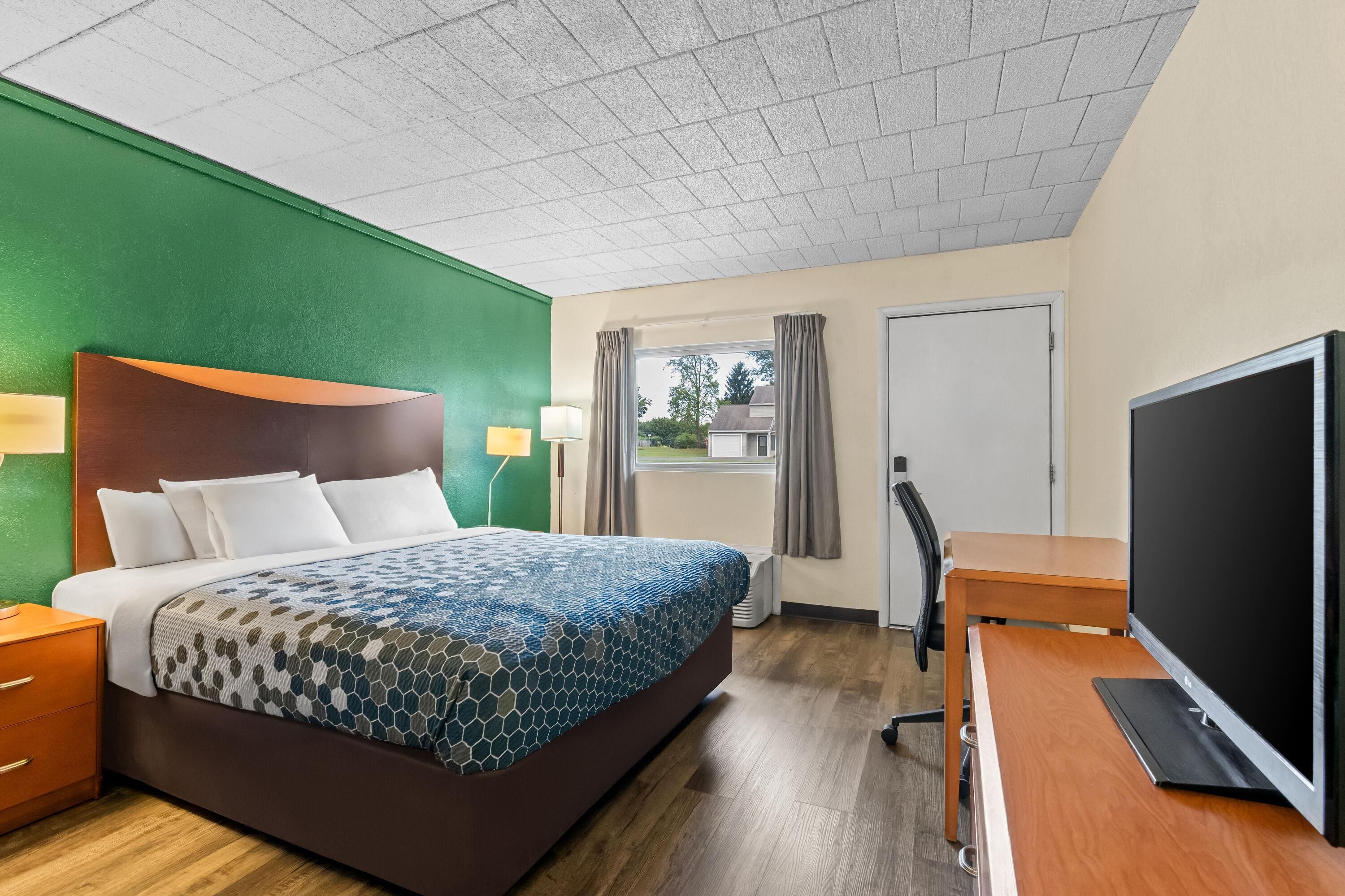 A hotel room with a bed, a desk with a TV, a chair, and a window with curtains.