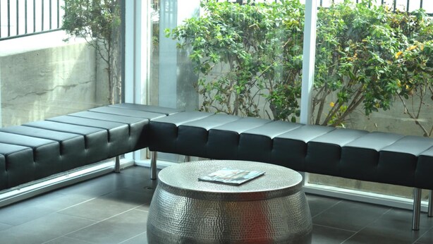 A modern seating area with a bench and a round table, a concrete wall, and a view of greenery outside. - Rodeway Inn South San Francisco Airport (South San Francisco)