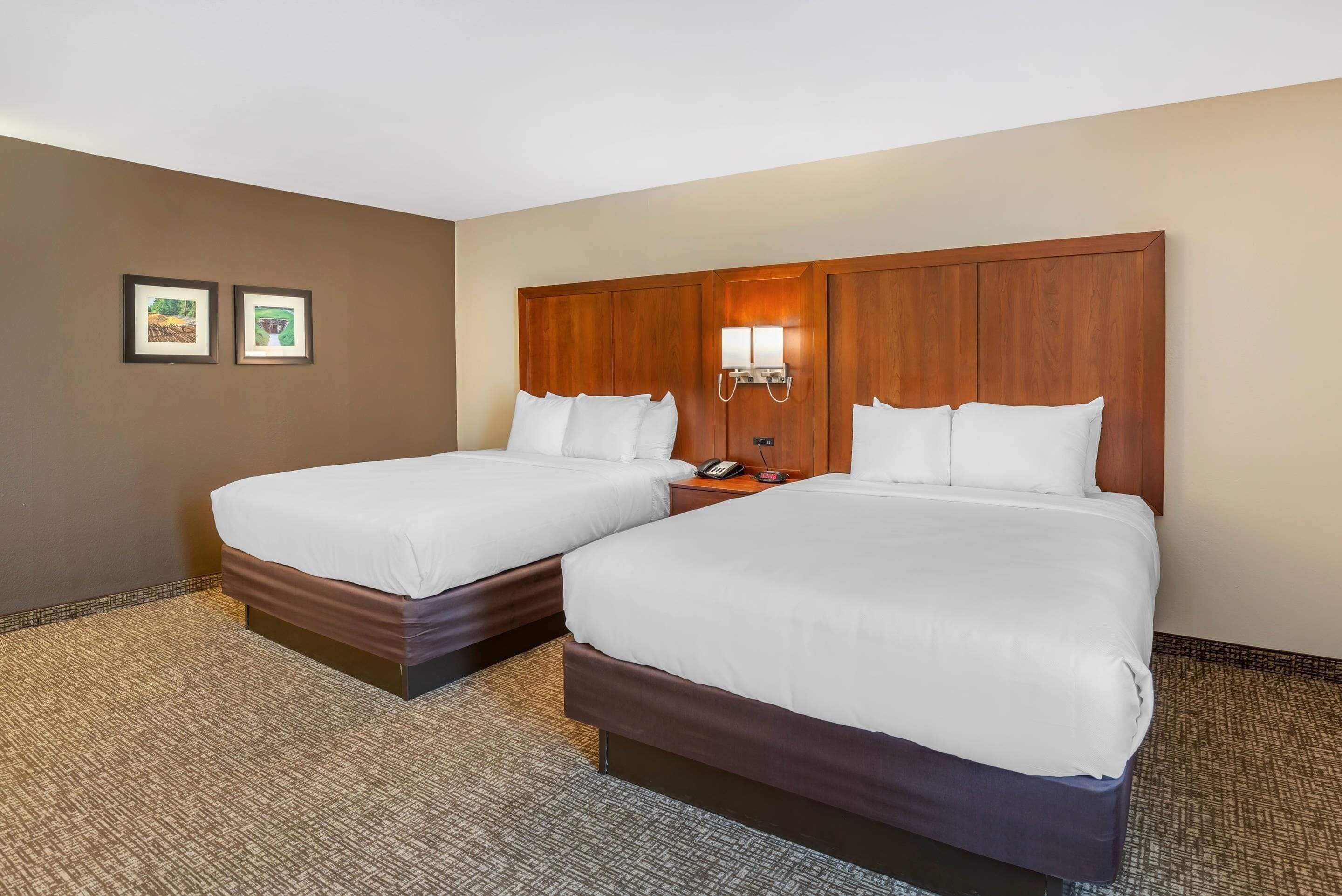 A hotel room with two beds, a wooden headboard, a nightstand with a phone, and two framed pictures on the wall.