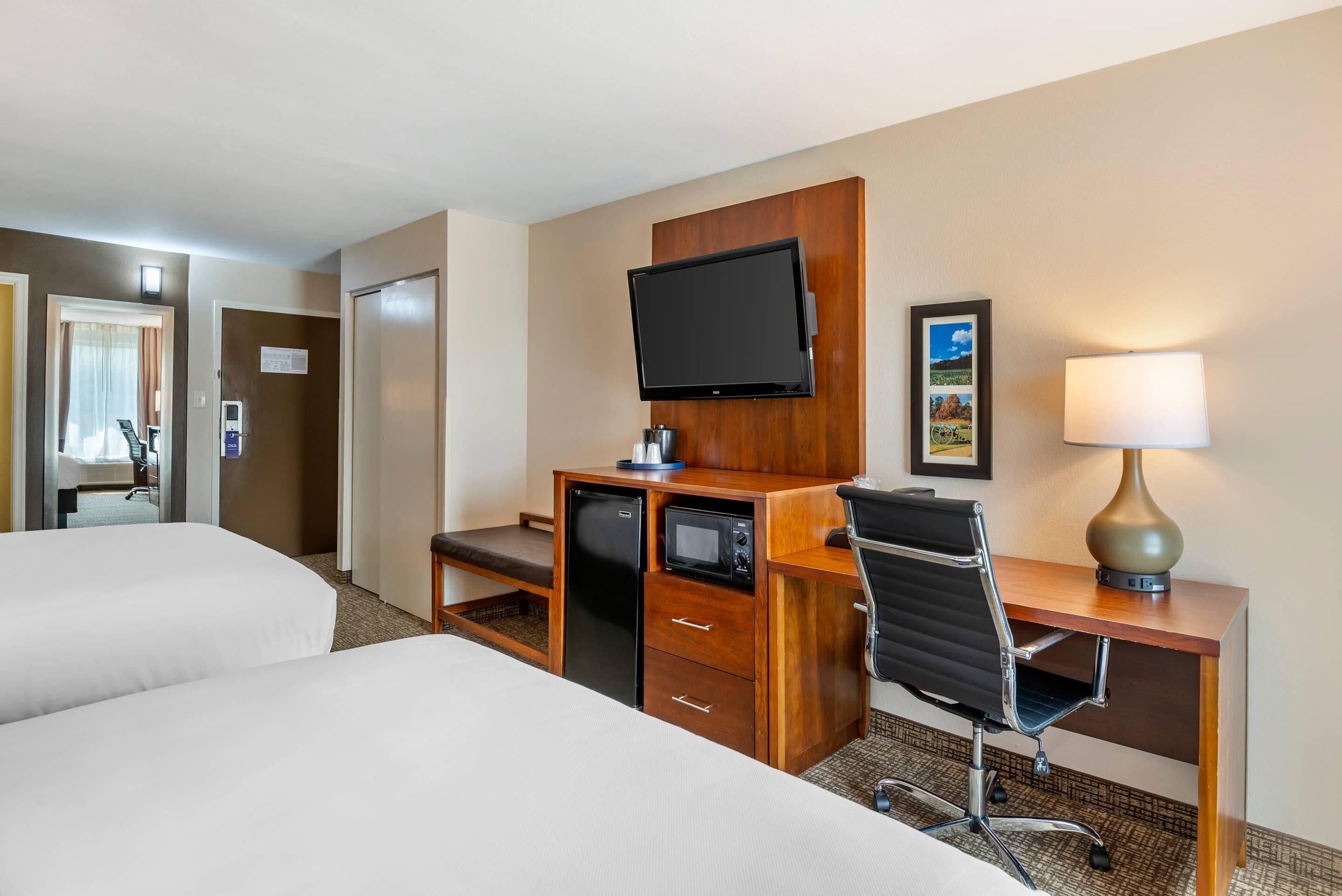 A hotel room with two beds, a desk with a television, a lamp, and a small refrigerator.