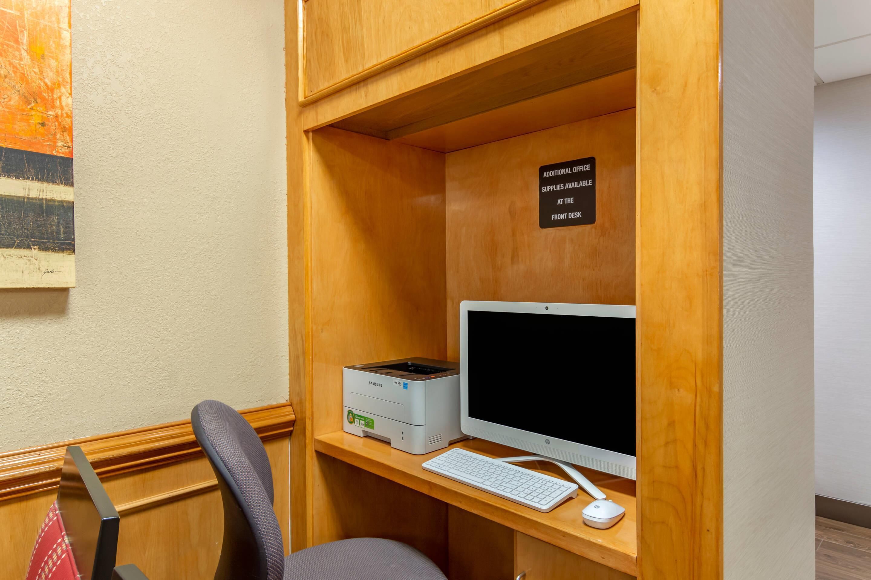 Un pequeño espacio de oficina con un escritorio, una computadora, un teclado y una silla. Hay un letrero en la pared que indica que hay útiles de oficina adicionales disponibles en la recepción.