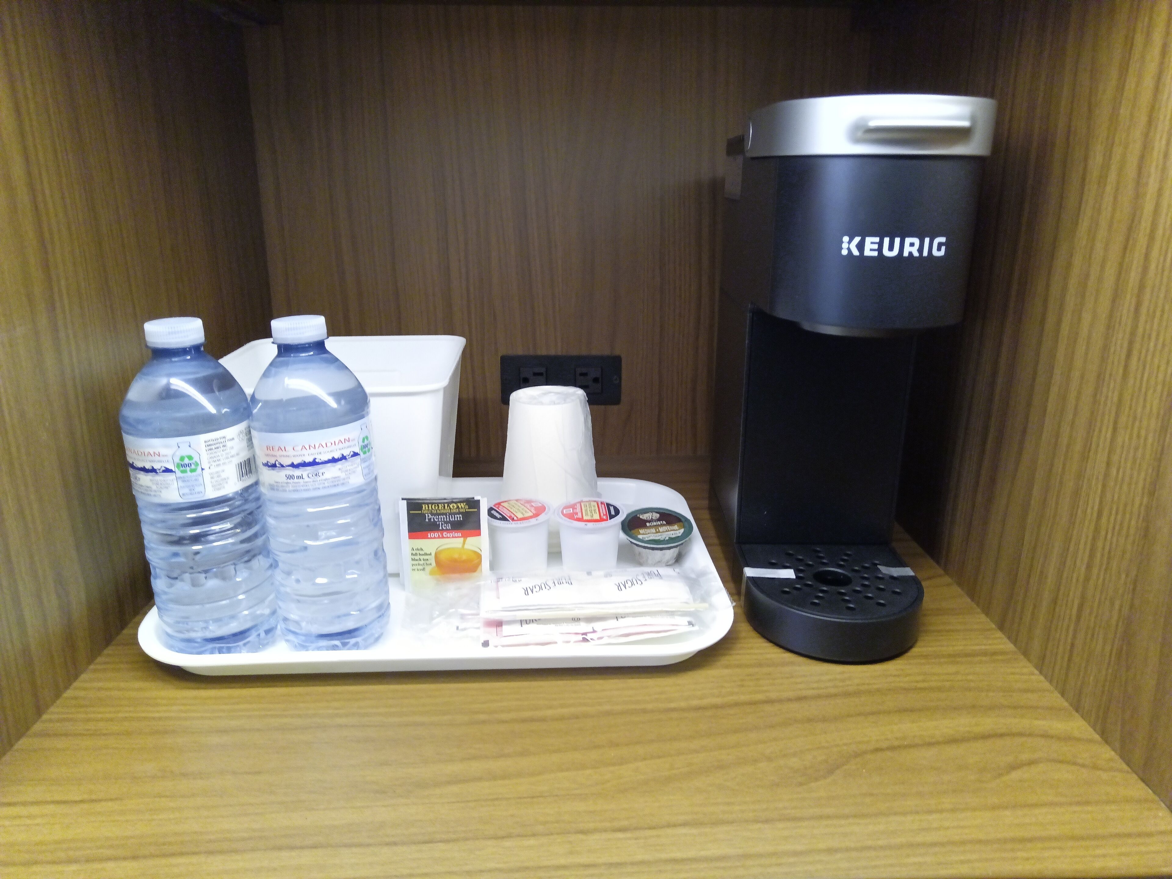 Una bandeja con dos botellas de agua, una cafetera, una taza, una caja de té y un pequeño recipiente.