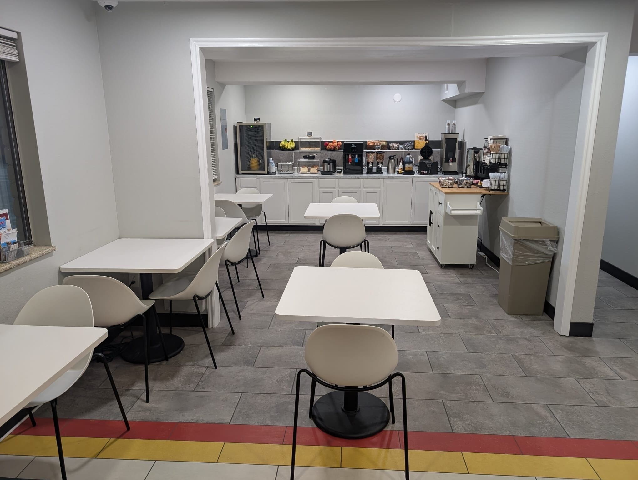 A clean, modern dining area with white tables and chairs, a kitchen in the background, and a trash bin to the right.