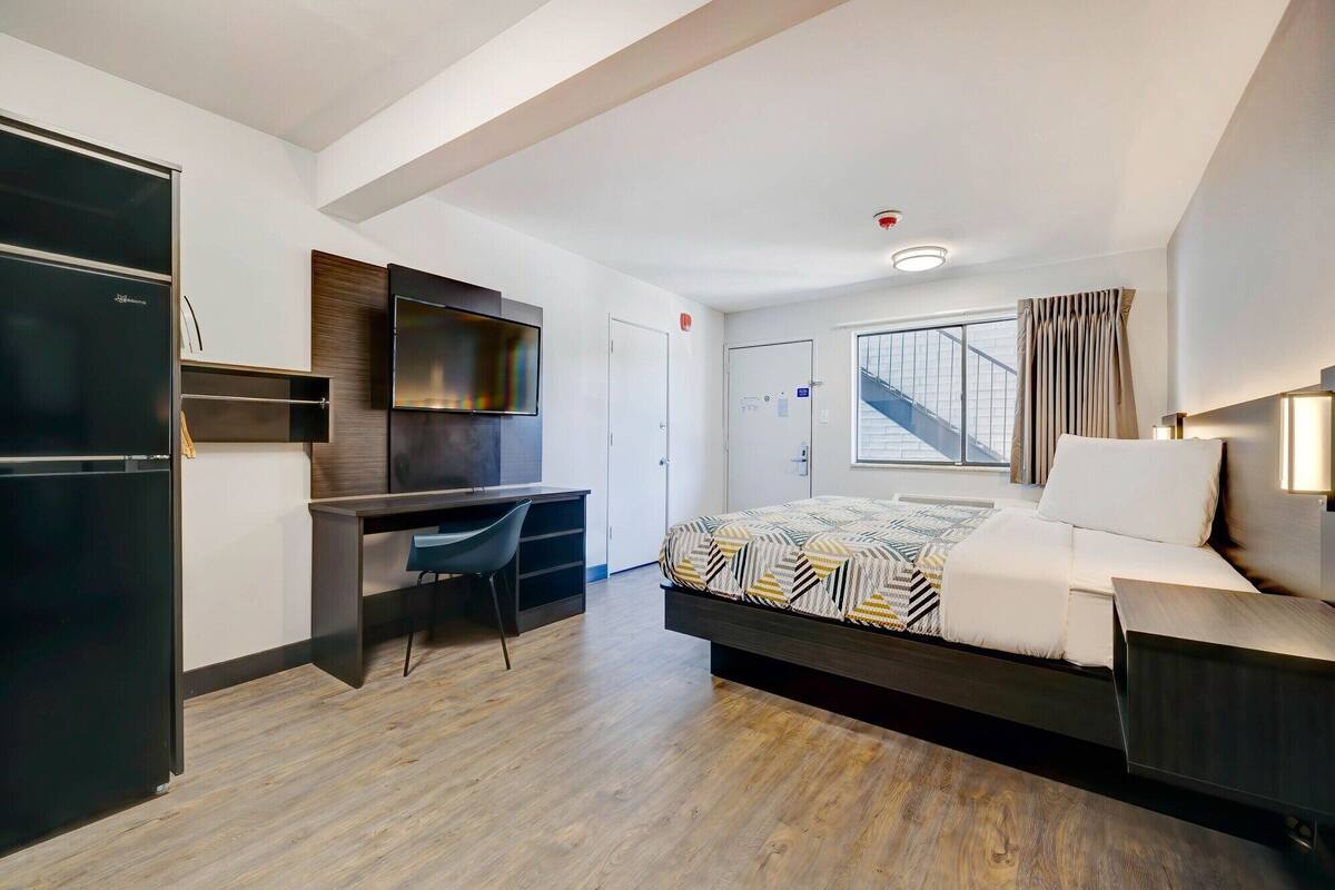 A modern hotel room with a bed, desk, chair, and a TV mounted on the wall.