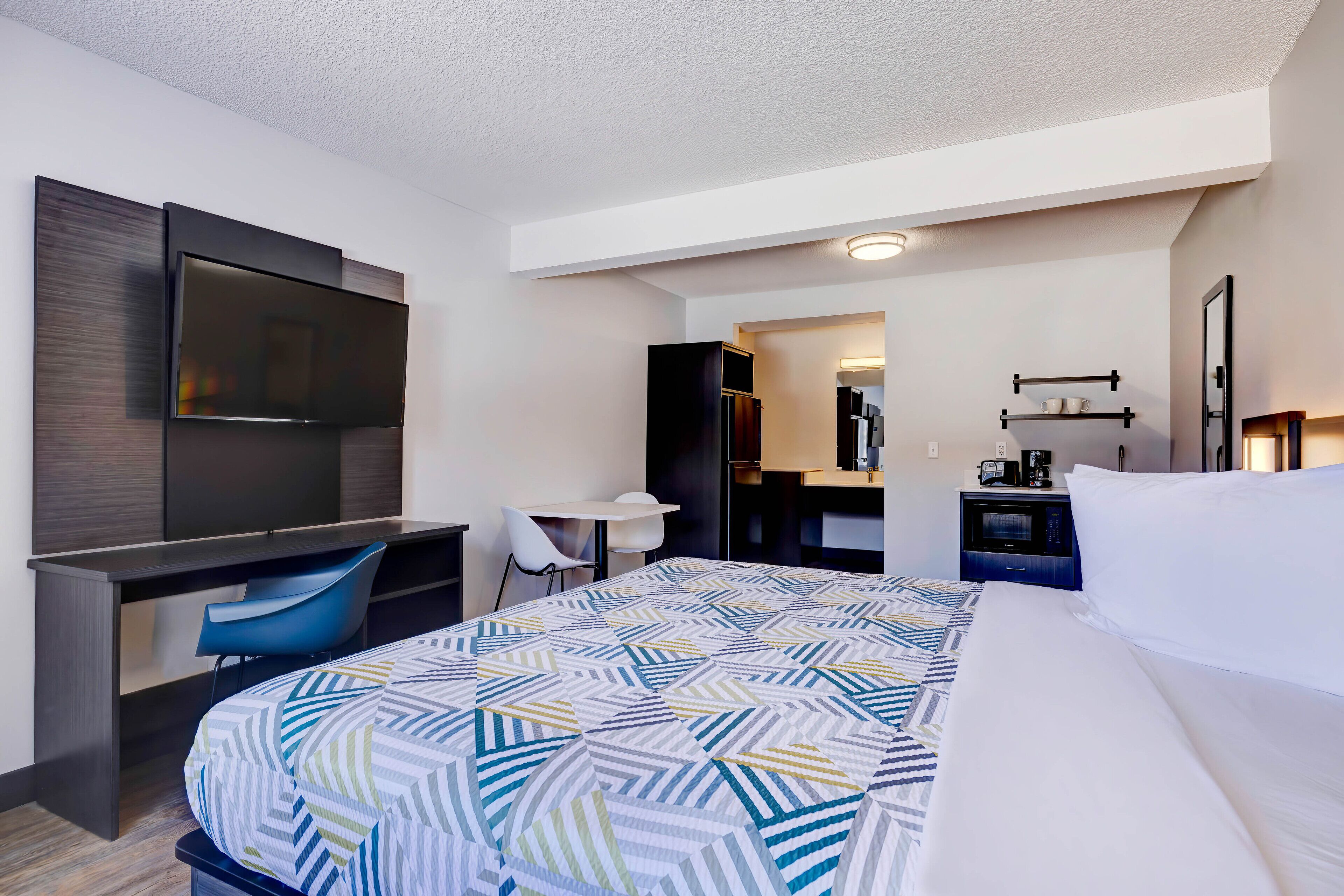 A hotel room with a large bed, a desk with a chair, a TV mounted on the wall, a small table with a lamp, and a microwave.