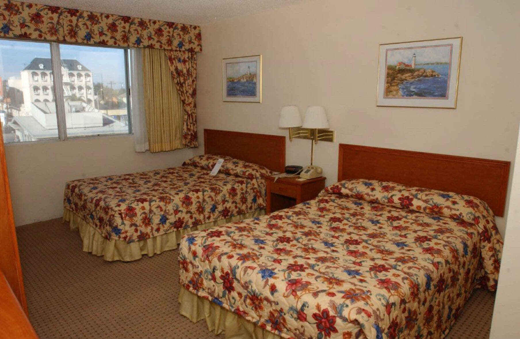 Une chambre d’hôtel avec deux lits, des couvre-lits à motifs floraux, une fenêtre avec des rideaux, une table de chevet avec une lampe et deux tableaux encadrés accrochés au mur.