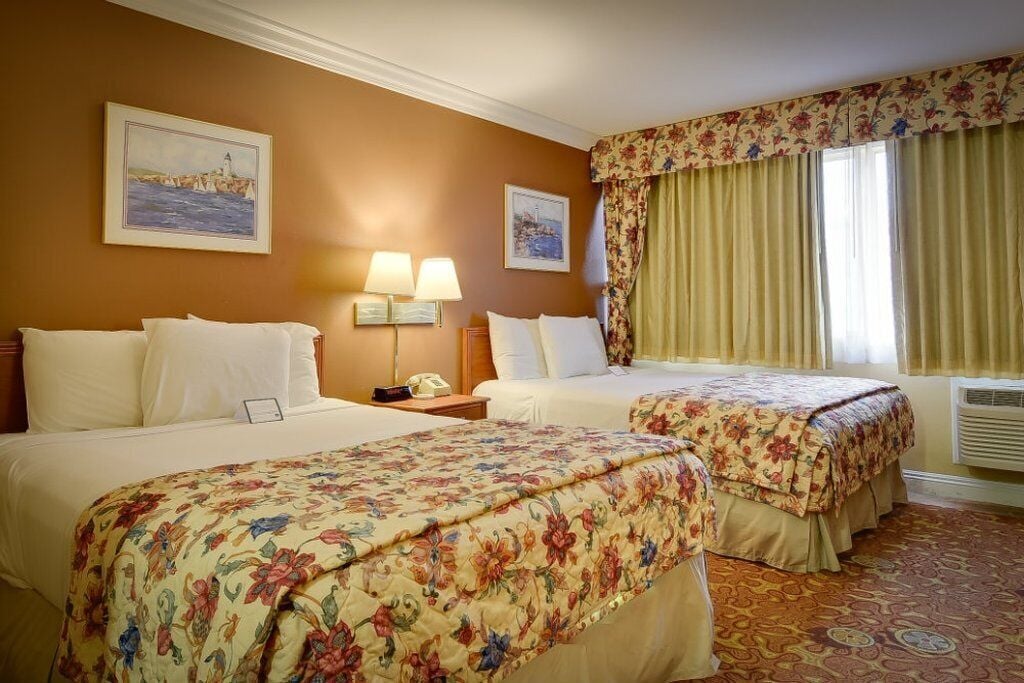 Habitación de hotel con dos camas, cubrecamas estampados con motivos florales, una mesita de noche con lámpara y un cuadro de un faro en la pared.