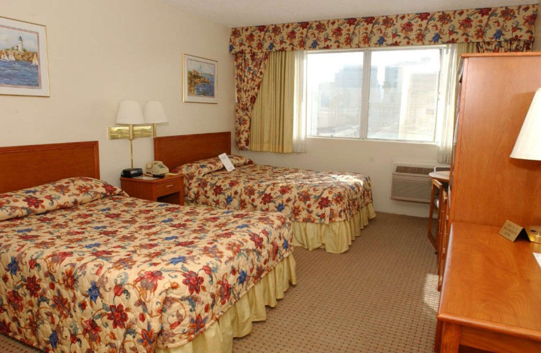A hotel room with two beds, a desk, a chair, a window with curtains, a lamp, and a painting on the wall.