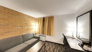 Una habitación de hotel con pared de ladrillo, un sofá, un escritorio con silla, un espejo y una ventana con cortinas.