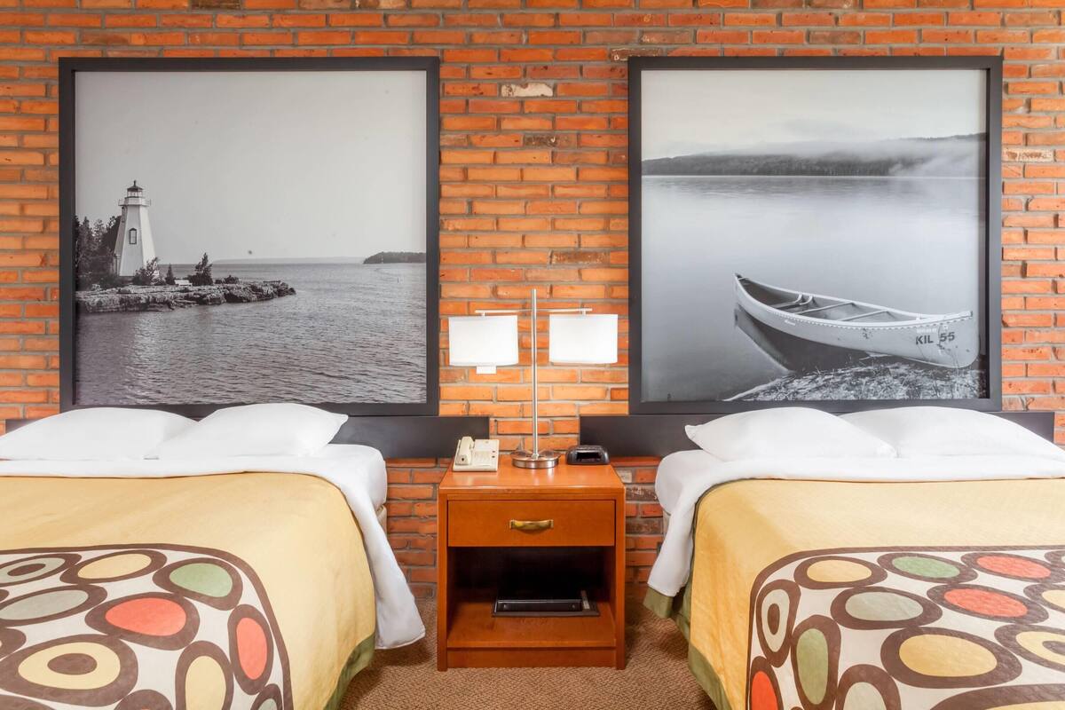 Une chambre d’hôtel avec deux lits, une table de chevet avec une lampe et deux tableaux représentant des paysages côtiers.